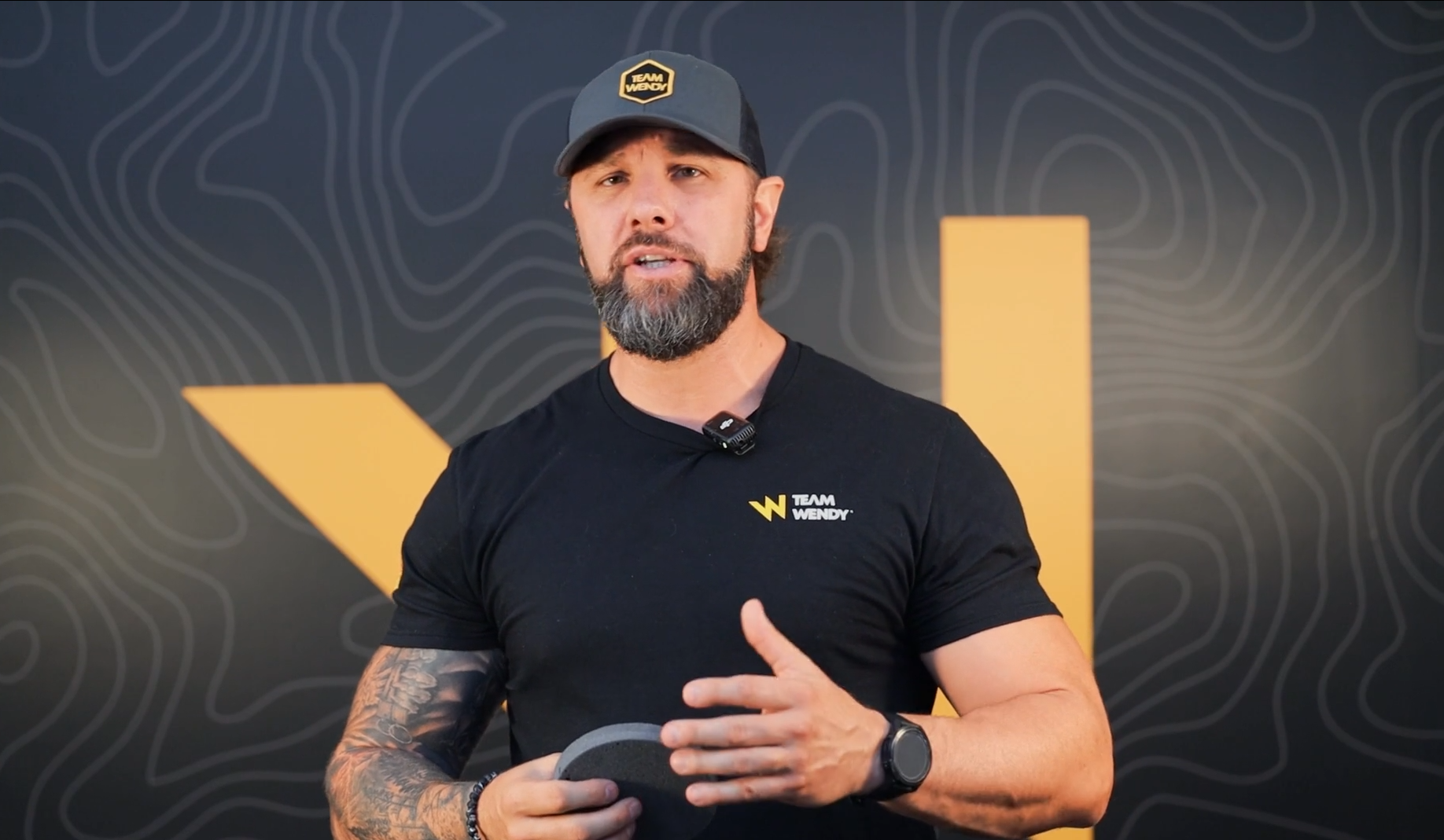 Man with beard and tattoos wearing a black team wendy tshirt and hat with a yellow w in the background