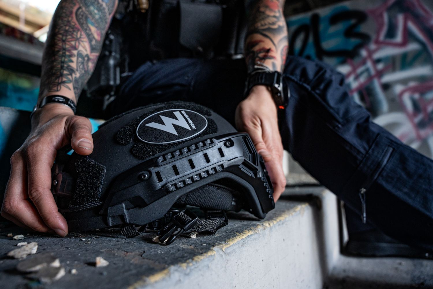 A law enforcement officer with tattooed arms picks up his black Team Wendy RIFLETECH rifle-rated ballistic helmet off concrete steps.