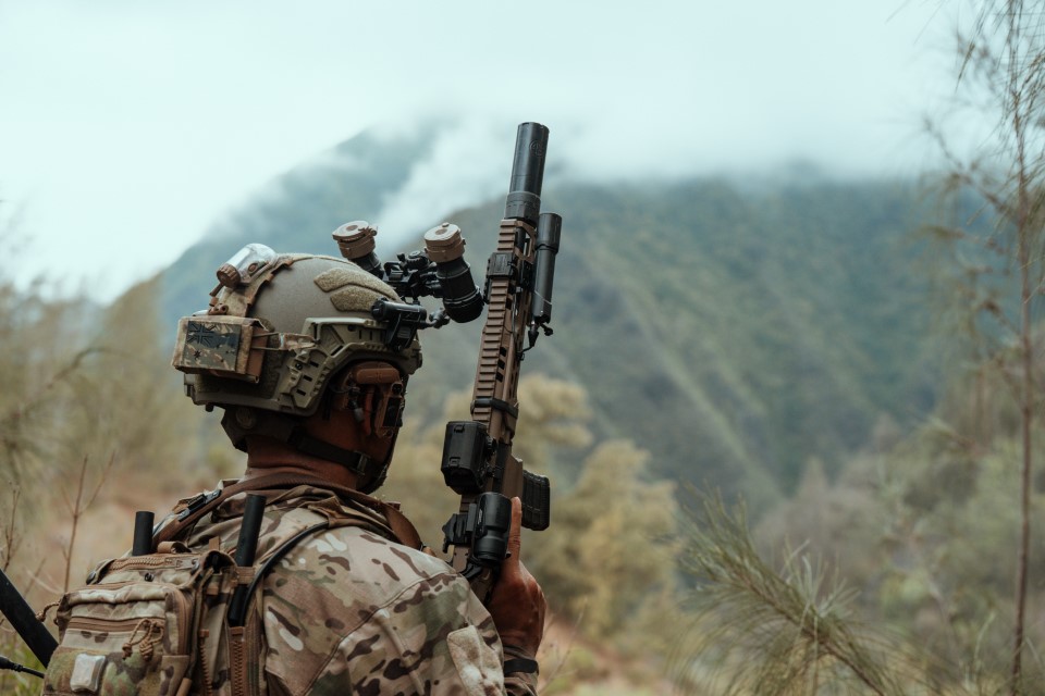 A soldier in camouflage gear wearing a Team Wendy RIFLETECH helmet stands outdoors holding a rifle with a suppressor, facing a mist-covered mountain in a dense, green forest setting