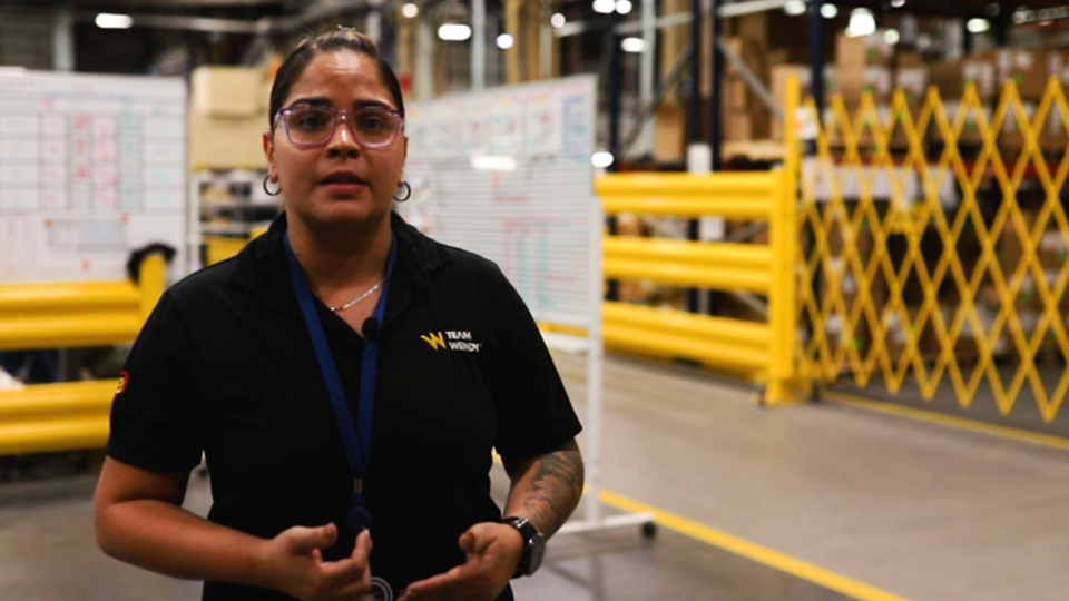 Woman wearing glasses, earrings and a necklace in a black team wendy t shirt inside of a warehouse