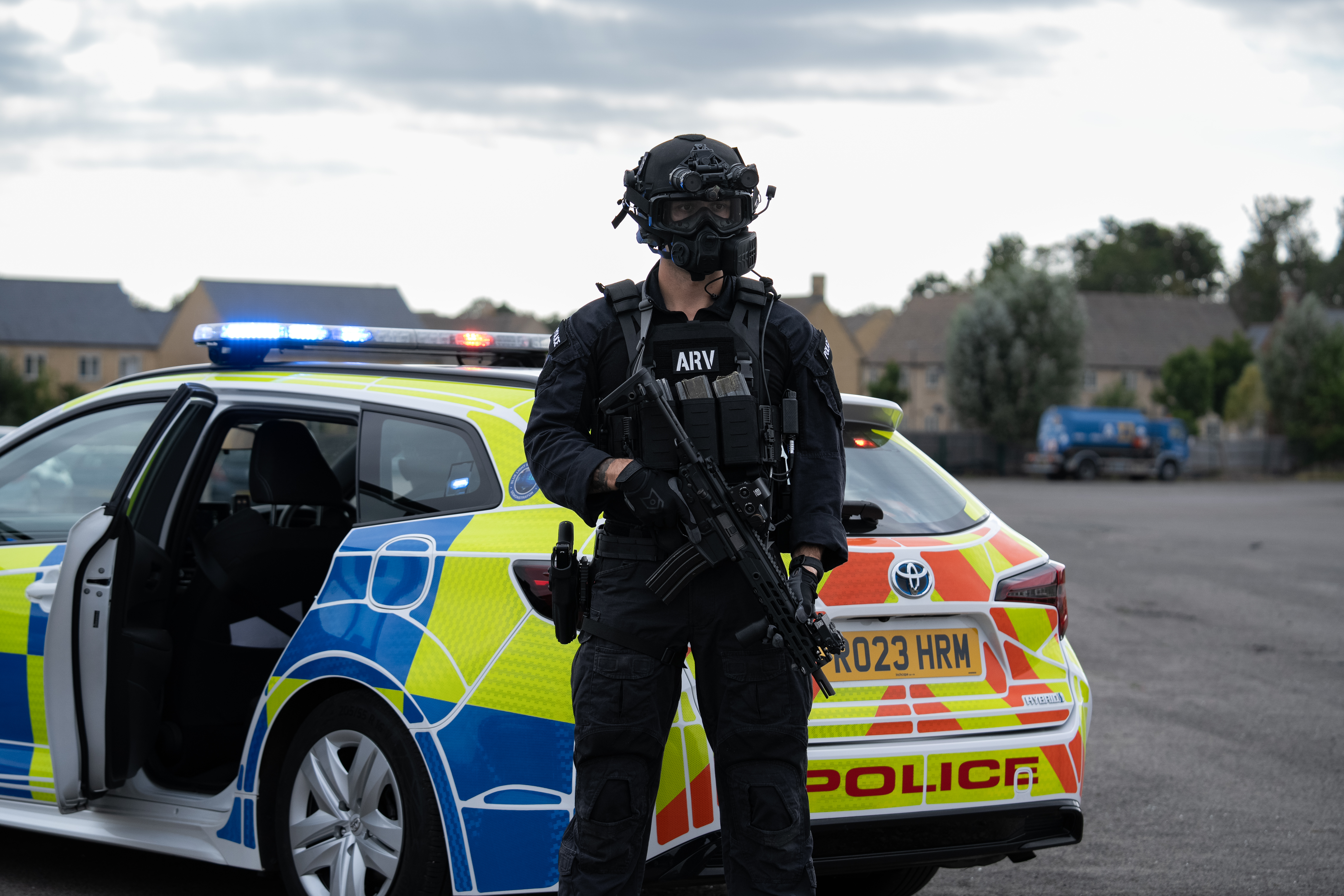 Police officer wearing all black with a gun, goggles and black Team Wendy helmet in front of a police vehicle