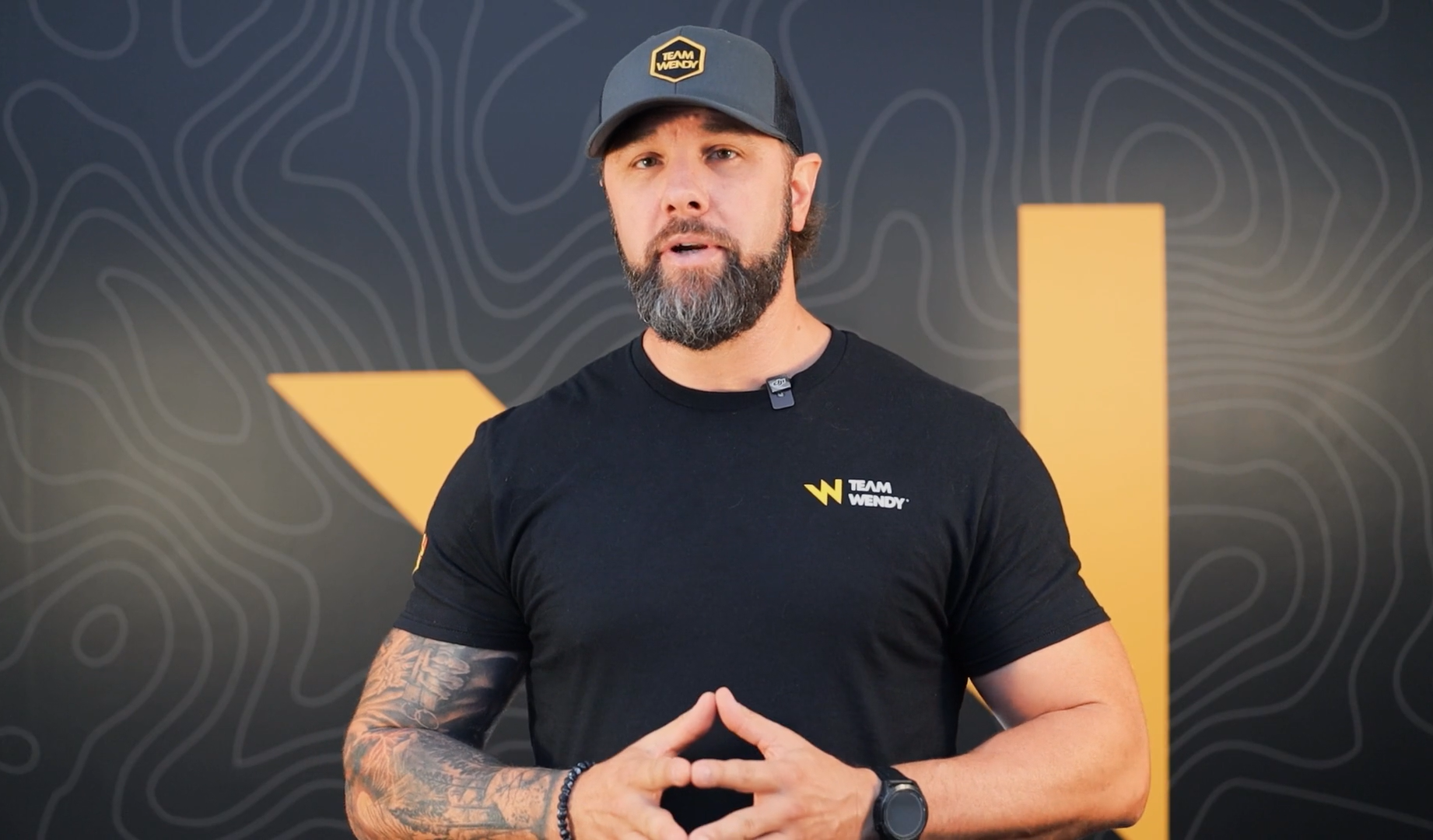 Image of man with beard and tattoos with a black Team Wendy tshirt and hat with a yellow W in the background