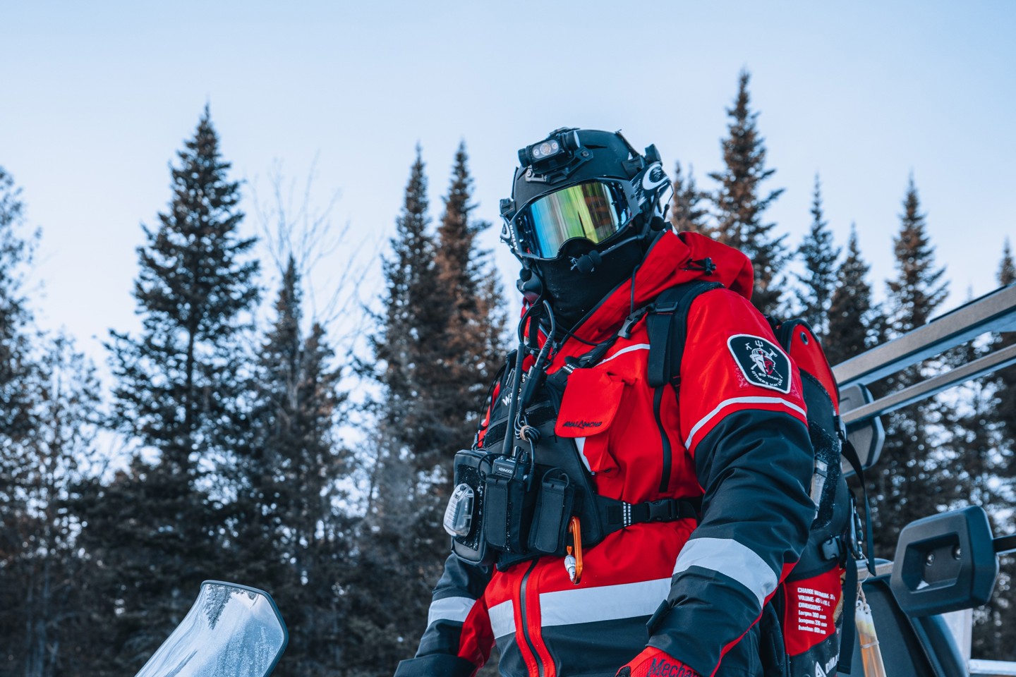 Person standing on a snow vehicle wearing a red jacket, goggles and a Team wendy helmet and radio rig with snow and trees in the background
